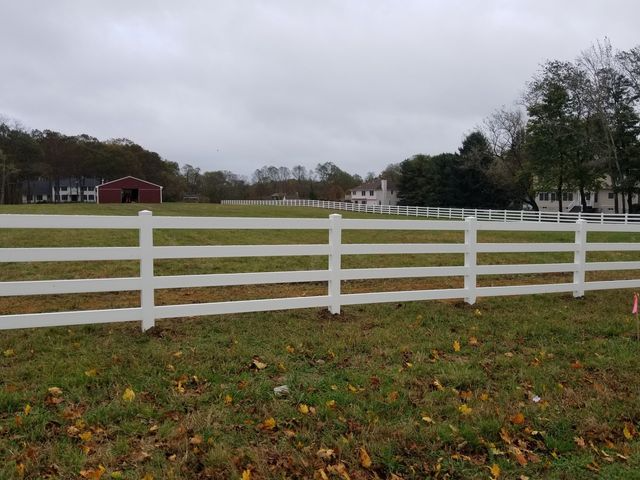 A white, four-rail fence stretches across a green grass field toward a red barn and houses under an overcast sky.