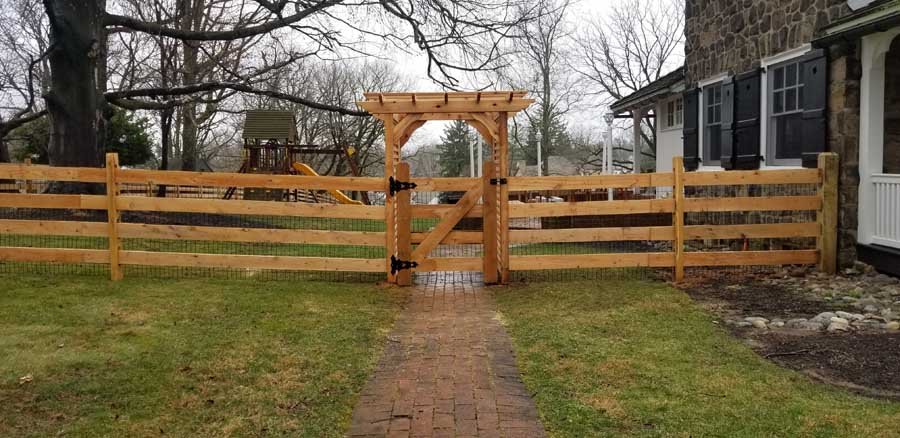 A wooden fence with a matching arbor and gate spans a lawn, with a brick path leading to the gate.