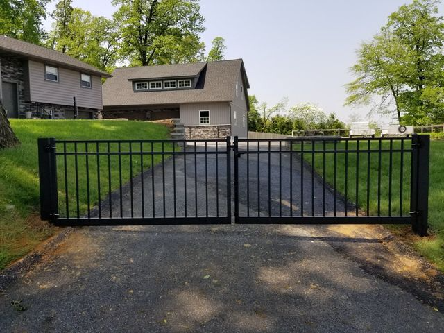 Black metal double gate across an asphalt driveway leading to a suburban house with green lawns and trees.