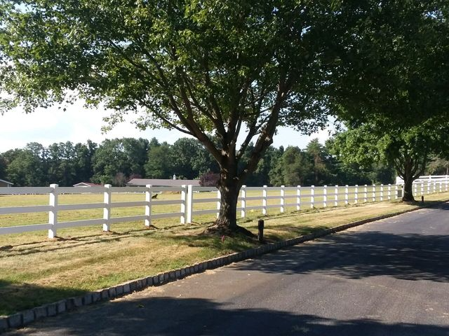 A white rail fence runs along a paved road next to a grassy field with trees under a clear sky.