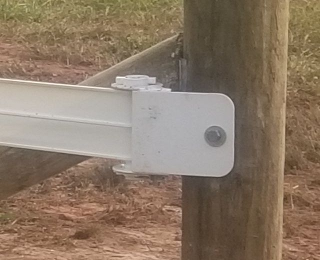 A white metal gate hinge is bolted to a wooden fence post in an outdoor setting.