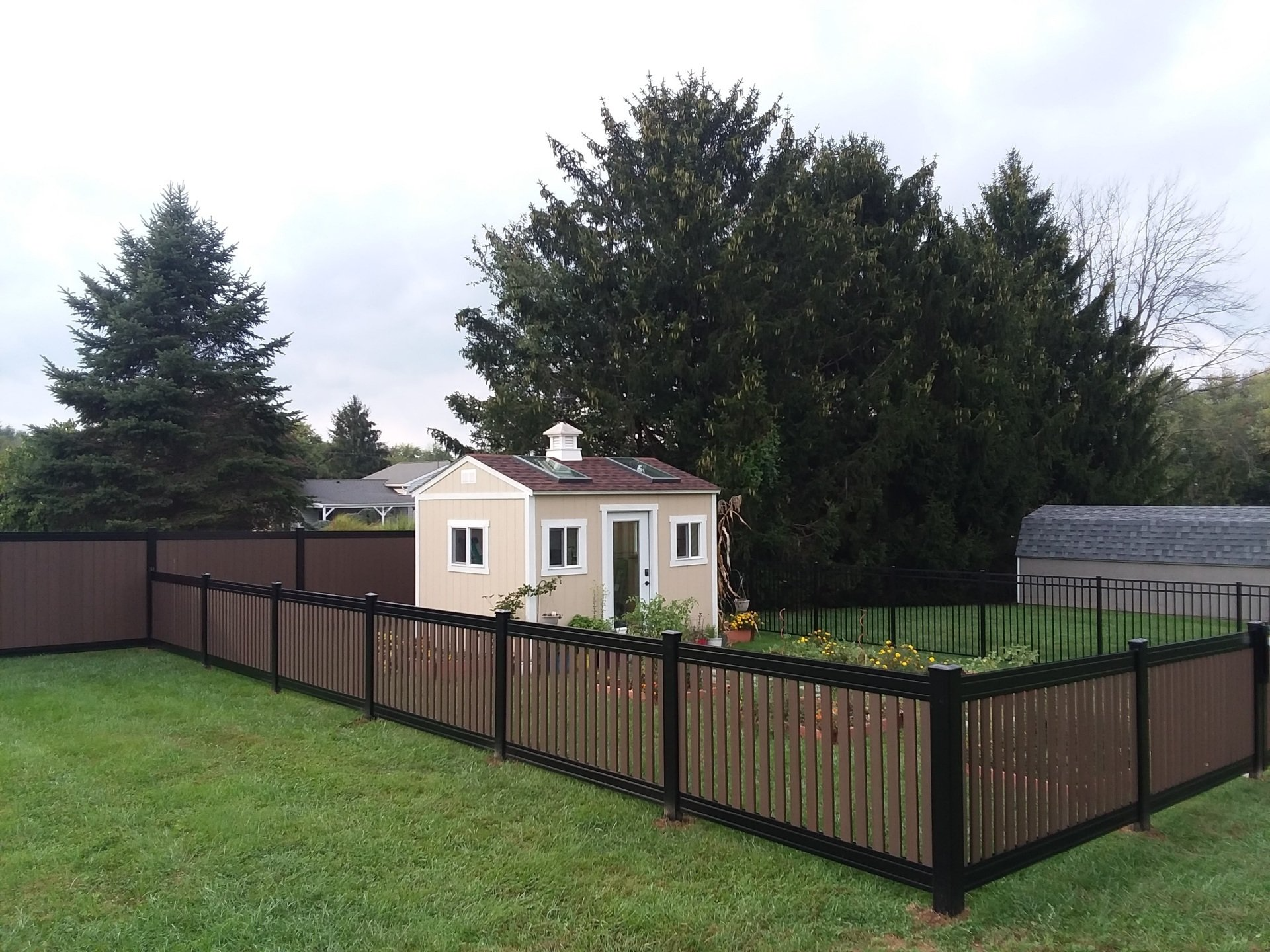 A tan shed with a small chimney sits in a backyard enclosed by a brown and black fence on a grassy lawn.