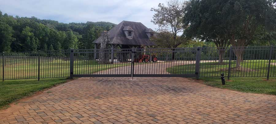 A dark metal gate stands at the entrance of a paved driveway leading to a stone pavilion in a green, tree-lined landscape.