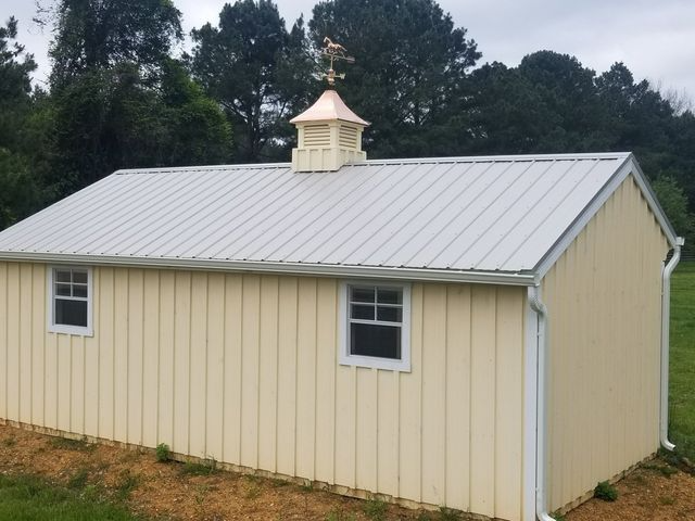 A light yellow shed with a metal roof, a white cupola with a weather vane, and two windows in a grassy setting.