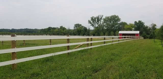 A long white fence stretches across a green grassy field leading toward a small red barn under a cloudy sky.