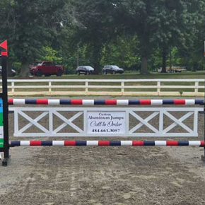 A horse show jump painted white with red and navy blue rails sits in a dirt arena. A sign reads