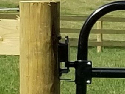 A black metal gate hinge mounted on a wooden fence post in a grassy field.