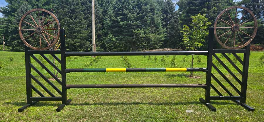 A horse jumping obstacle with black metal stands, decorative wagon wheels at the top, and two green and yellow rails.