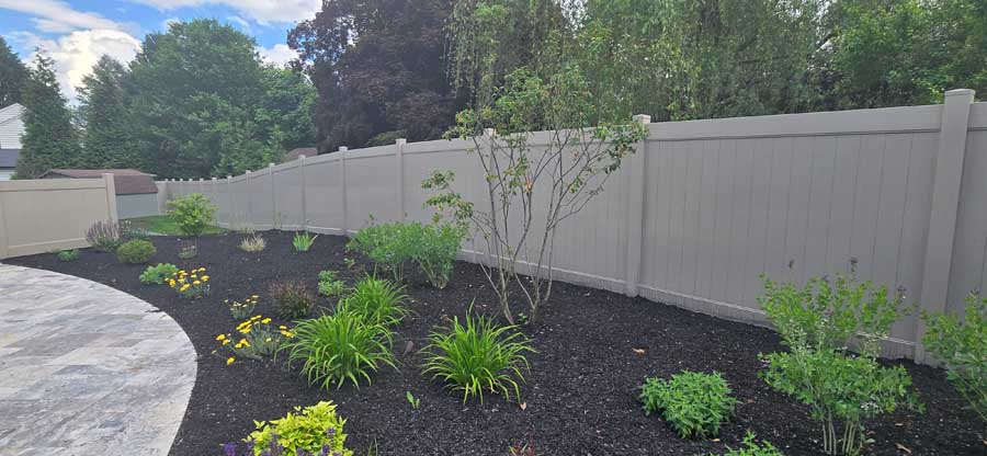 A garden bed with young shrubs and flowers mulched in dark soil, bordered by a stone patio and a tan vinyl privacy fence.