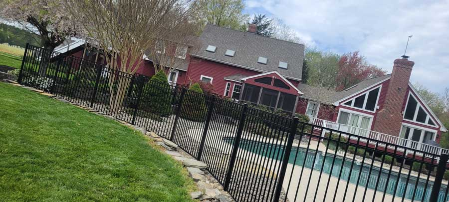 A red two-story house with a backyard pool enclosed by a black metal fence on a grassy lawn under a cloudy sky.