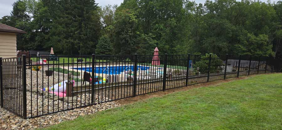 A backyard swimming pool enclosed by a black metal fence, bordered by gravel and grass with dense trees in the background.