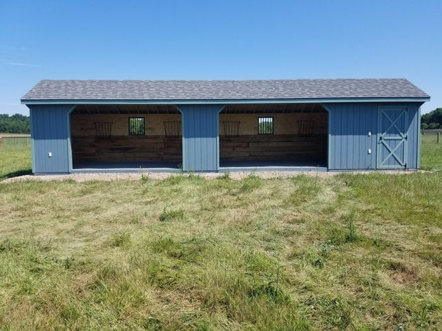 A blue, two-stall run-in shed with a shingled roof and a tack room door, situated in a grassy field.