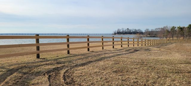 A long wooden fence runs across a dry, grassy field toward a calm lake under a clear, bright sky.