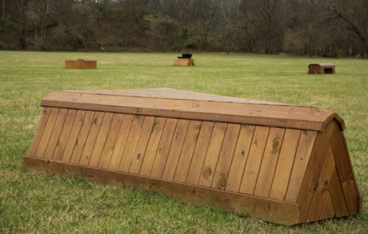 A wooden wedge-shaped jump for horse cross-country training sits on a grassy field with other jumps in the background.