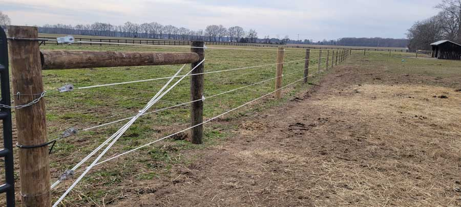 A wooden fence line with wire and rope segments separating a grassy field from a dirt patch under a cloudy sky.