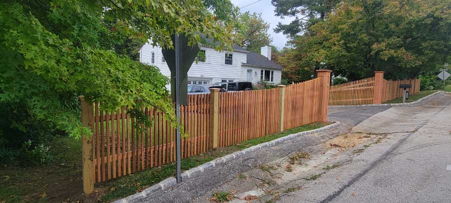 New natural wood picket fence lining the front of a suburban property next to an asphalt road and sidewalk.