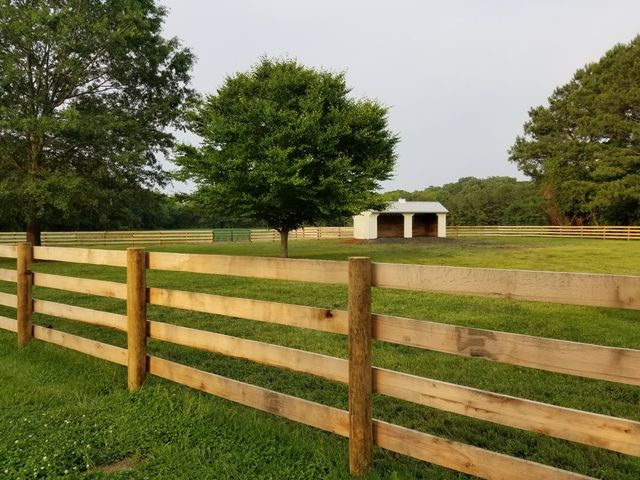 A wooden four-rail fence borders a green pasture with a small white shed and trees under a clear sky.