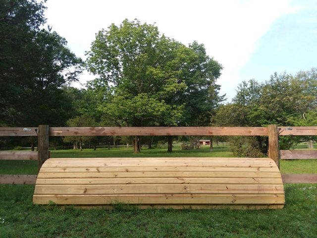 A wooden cross-country equestrian jump, rounded and plank-sided, set in a grassy field in front of a wooden fence.