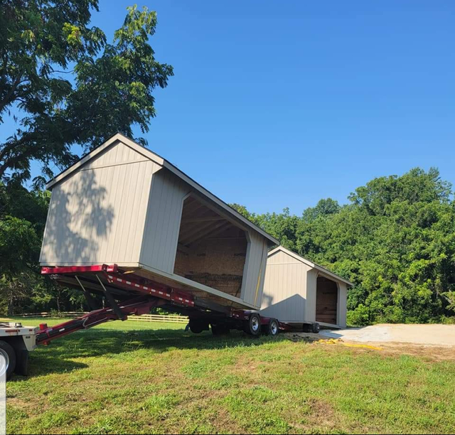 Two light-tan prefabricated sheds are being transported on a specialized flatbed trailer in a grassy, wooded area.