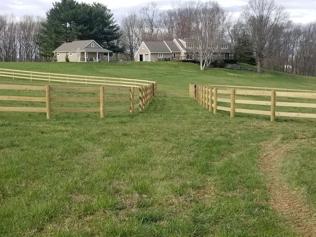 A wooden fence borders a grassy field with two houses and trees in the background under a cloudy sky.