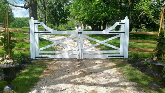 A white wooden farmhouse-style driveway gate with black hinges, set against a gravel road and green trees.