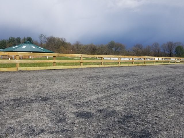 A gravel equestrian arena bordered by a wooden post-and-rail fence, with a green structure and trees in the background.