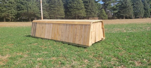 A wooden, trapezoidal horse jump obstacle sits in a grassy field in front of a tree line.
