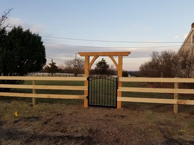 A black metal gate set within a natural wood arbor and a three-rail wooden fence on a rural property at sunset.