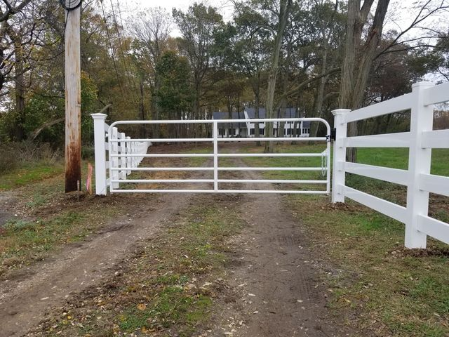 A white metal gate closed across a dirt driveway, flanked by white vinyl fencing in a grassy, wooded area.