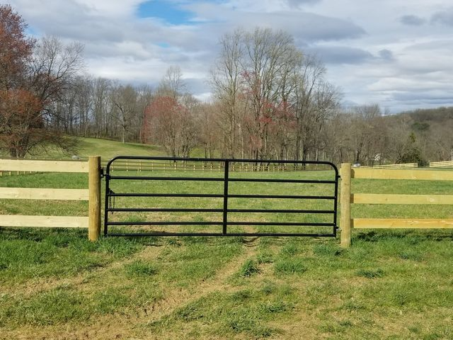 A black metal farm gate installed between two wooden fence posts in a grassy field with trees in the background.