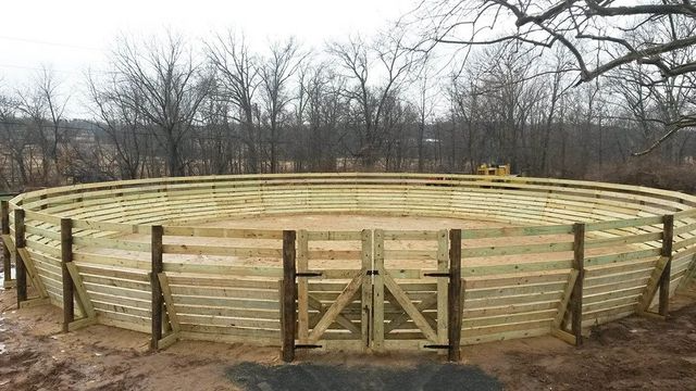 A round wooden horse training pen featuring a gated entrance, set outdoors against a backdrop of bare trees.
