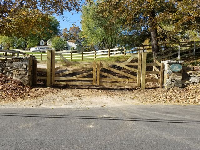 A wooden gate sits between two stone pillars at the entrance to a driveway, set against a backdrop of trees and a lawn.