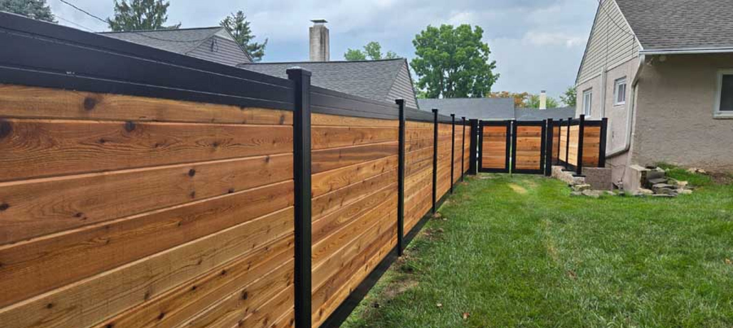 A modern horizontal wood fence with black metal posts and trim borders a residential backyard lawn.