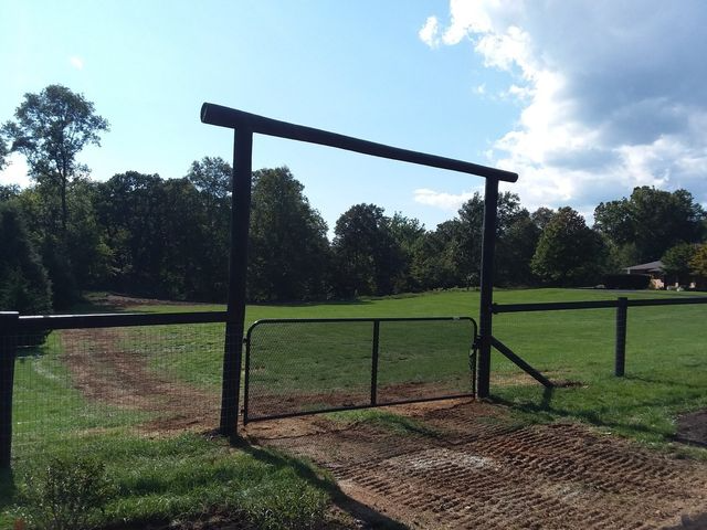 A metal gate set within a wooden frame on a rural property with a dirt path leading into a grassy field under a blue sky.