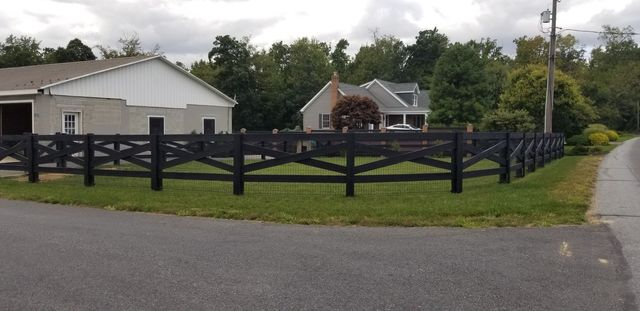 A dark wooden crossbuck fence borders a grass lawn with a white building and a house in the background.