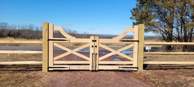 A pair of light-colored wooden gates closed between two fence posts on a gravel path against a blue sky.