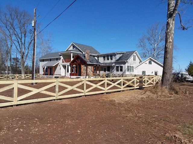 A light-colored two-story house with a stone porch and a new wooden X-brace fence in a rural, sunny setting.