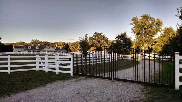 A gravel driveway enters through a tall black iron gate, surrounded by white rail fencing and a house in the distance.