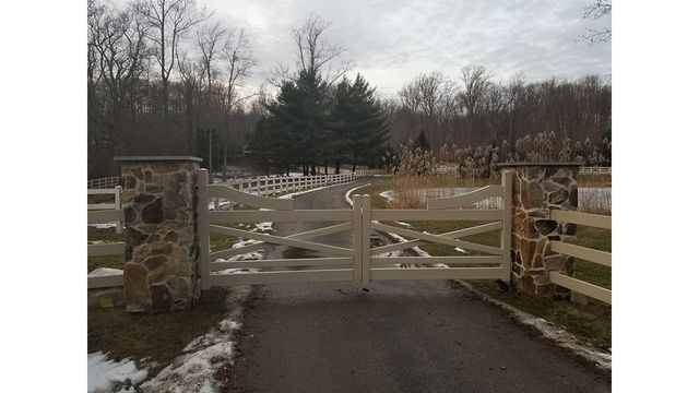 A closed, light-colored wooden gate set between two stone pillars, marking the entrance to a driveway near a treeline.