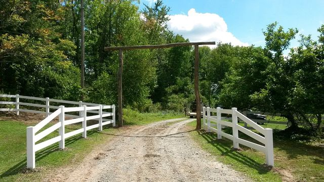 A white wooden fence lines a gravel driveway leading to a rustic wooden gate archway surrounded by lush green trees.