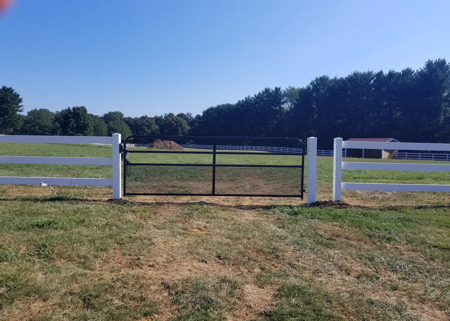 A black metal gate set between two white wooden fences in a grassy field under a clear blue sky.