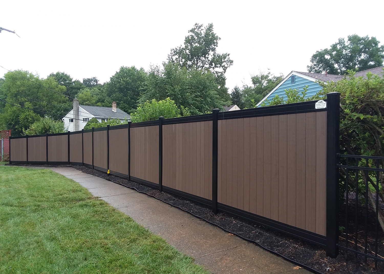A long privacy fence with brown vertical panels and black posts and trim, bordering a sidewalk and lawn.
