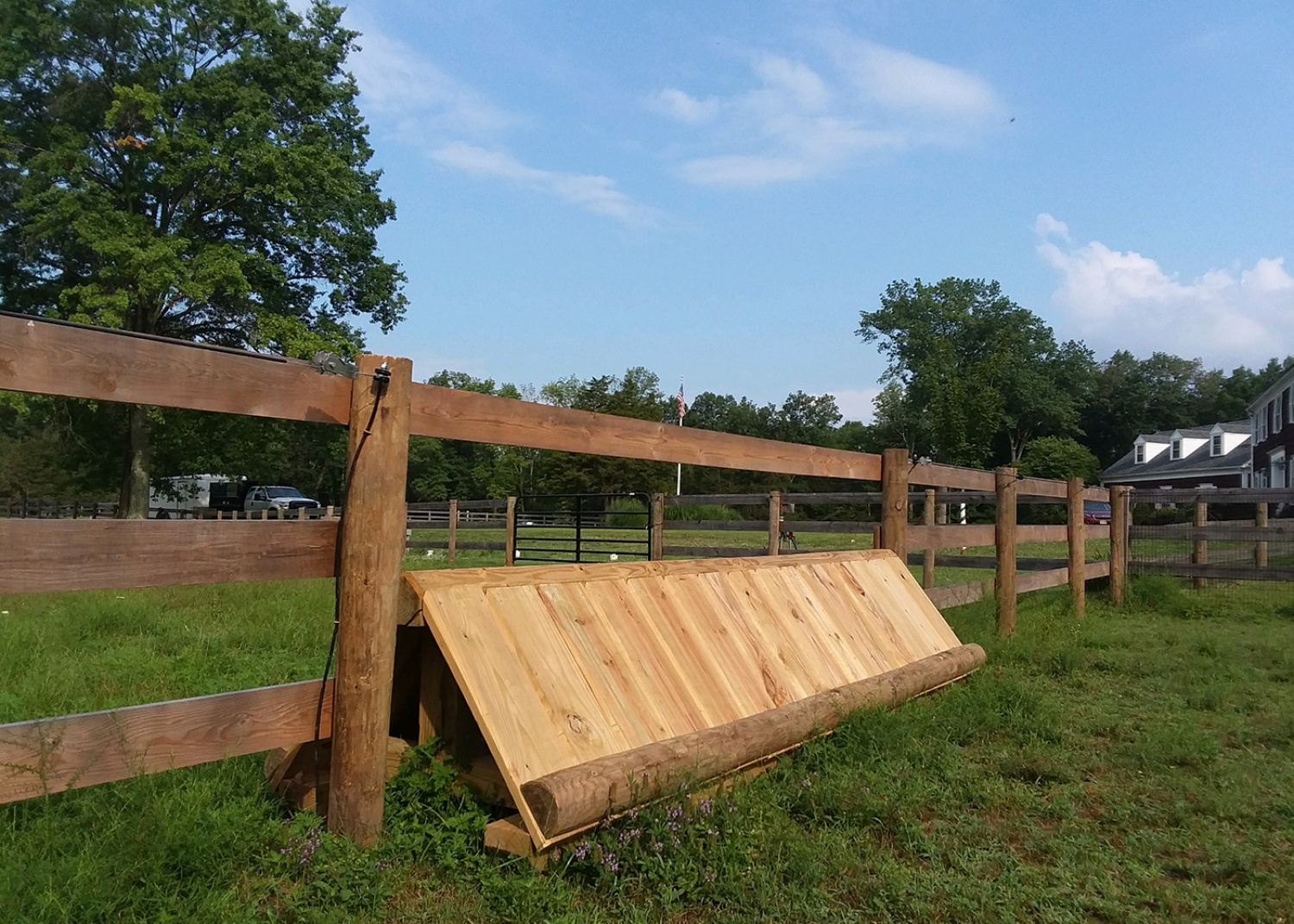 A wooden cross-country equestrian jump sits in a grassy field in front of a wooden rail fence.