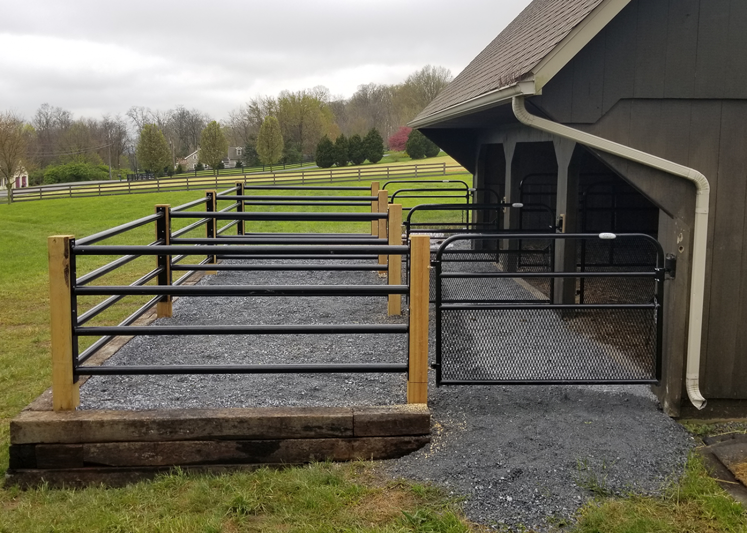 A gravel animal pen with black metal fencing attached to a wooden barn, set in a grassy rural landscape.