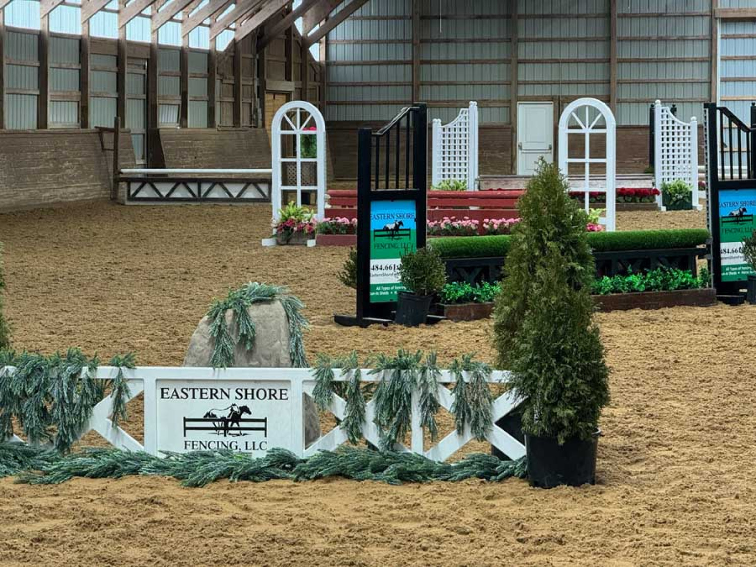 An indoor horse jumping arena featuring several white-arched jumps, decorative greenery, and a branded fence.