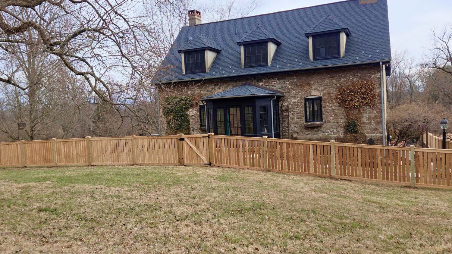 A two-story stone house with a slate roof and dormer windows, surrounded by a light-colored wooden picket fence.