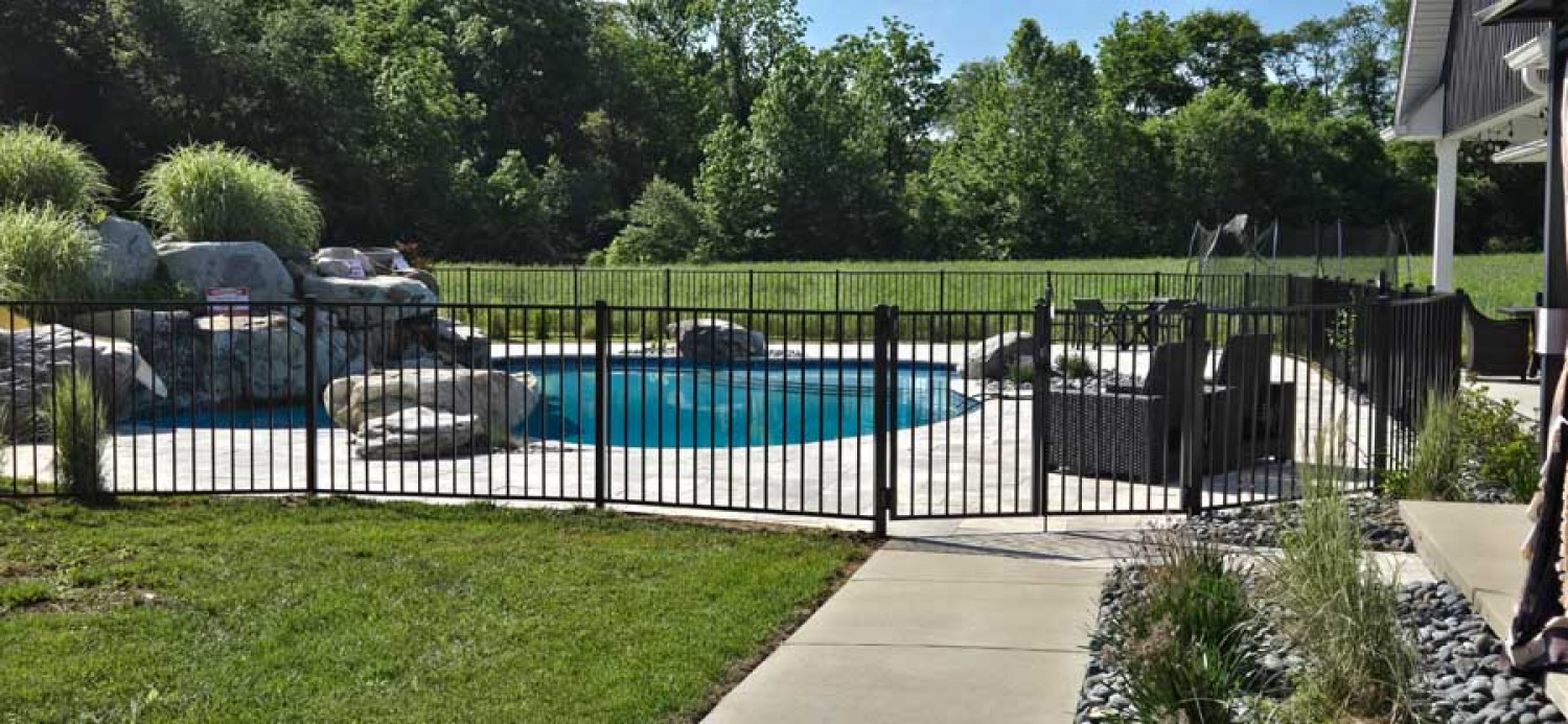A backyard swimming pool with a rock waterfall feature, enclosed by a black metal fence, next to a paved patio area.