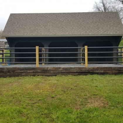A dark, three-bay open shed with a shingled roof, set behind a wooden retaining wall and a horizontal metal rail fence.