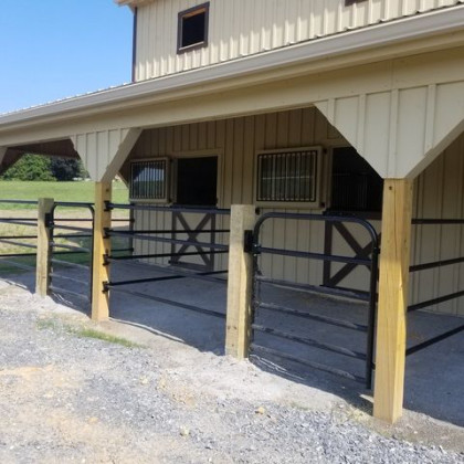 Tan barn exterior featuring two outdoor horse stalls with metal railings and gates under a covered porch.