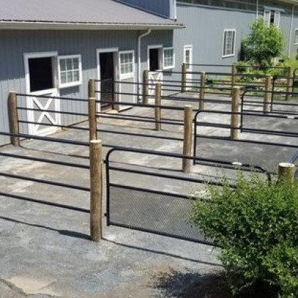 An outdoor area with gray barn stalls featuring metal gates and wooden posts on a paved lot.